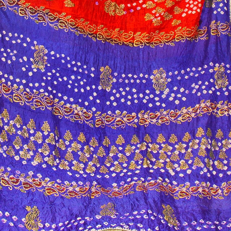 Close view of a blue and red silk bandhani dupatta covered in rows of small white tie-dyed dots, with gold paisley borders and figures worked over the dyed ground