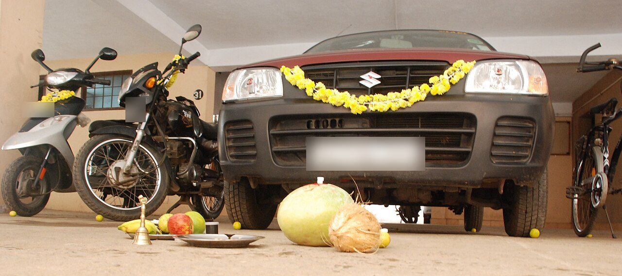 A car, a motorcycle and a scooter in a residential garage, each garlanded with yellow flowers for Ayudha Puja, with an ash gourd, a coconut, fruit, a bell and a puja plate set on the floor in front of them