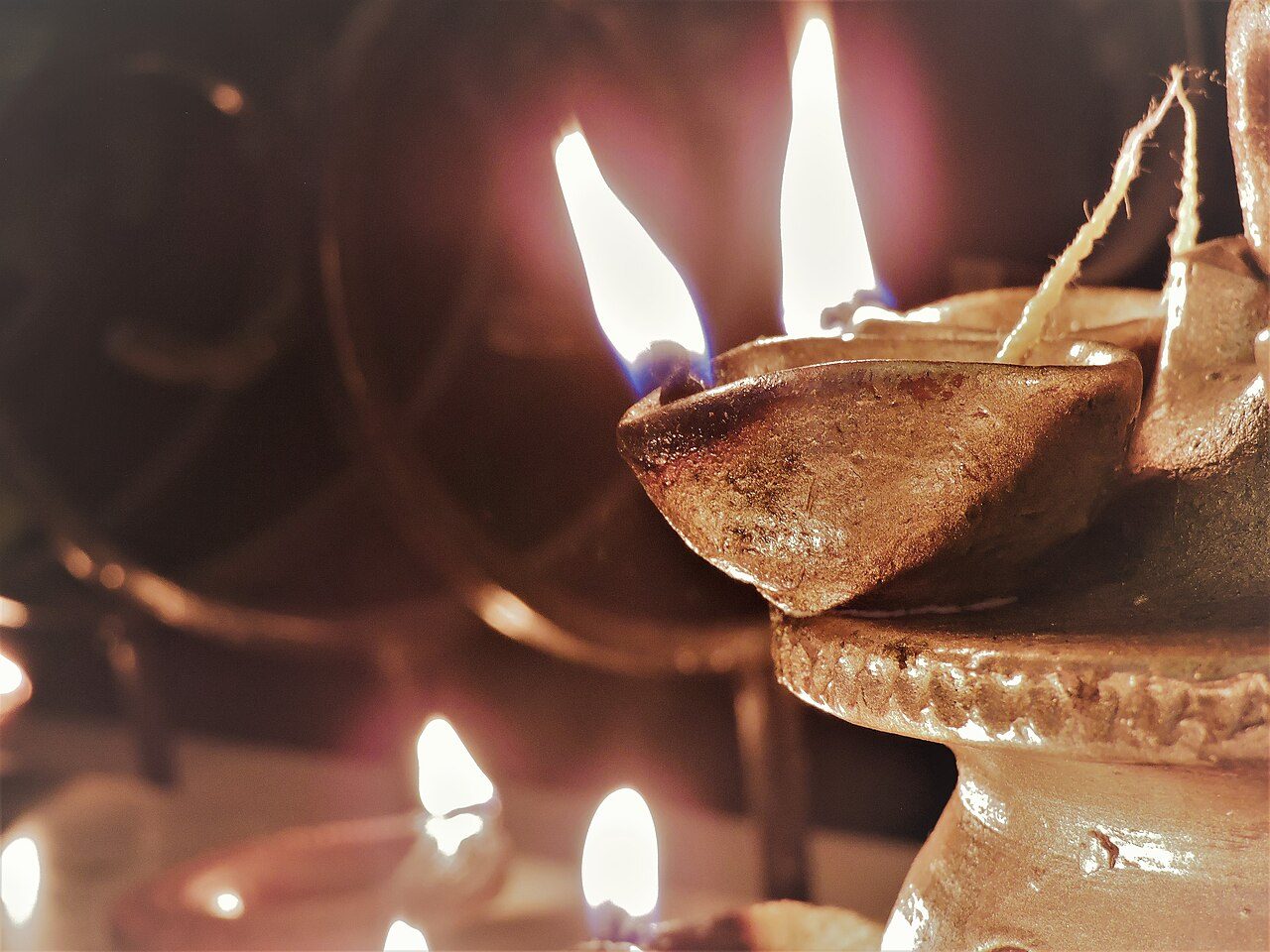 Close view of a hand-made clay diya with two burning cotton wicks, other lit diyas blurred in the background