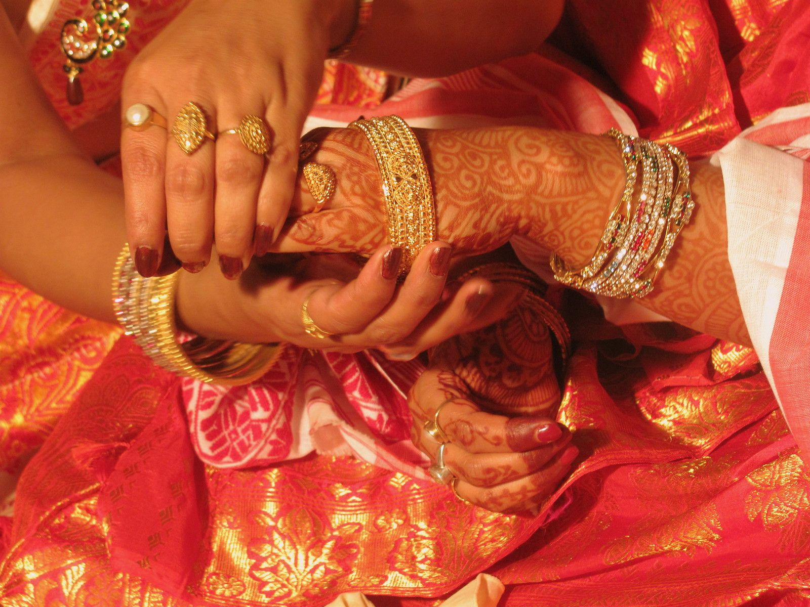 A woman wearing gold rings slides a carved gold bangle onto a bride's hennaed wrist at an Assamese juroon, over red and gold silk