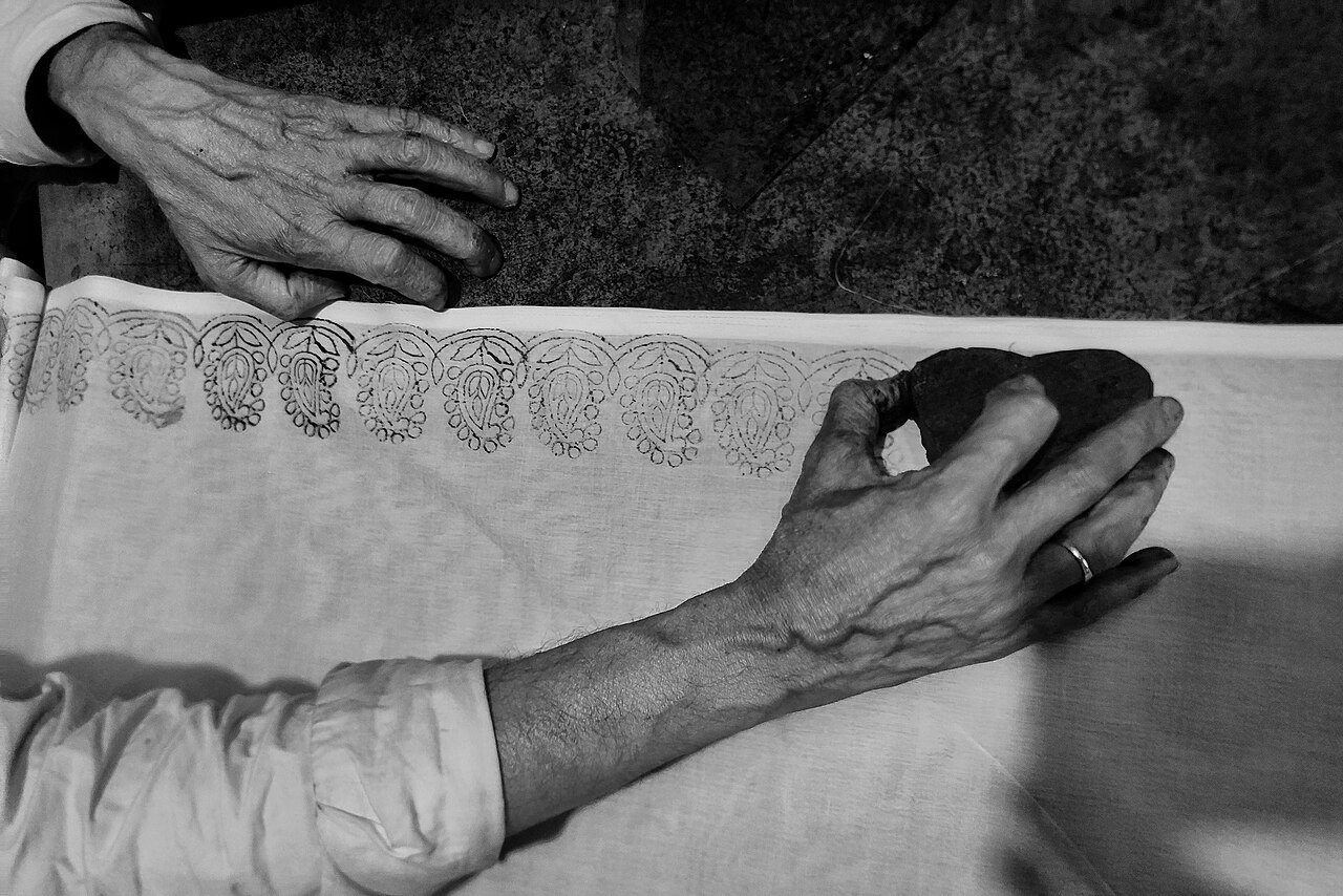 Close-up of a printer's hands pressing a carved block onto white cloth, leaving a row of printed motifs for chikankari embroidery