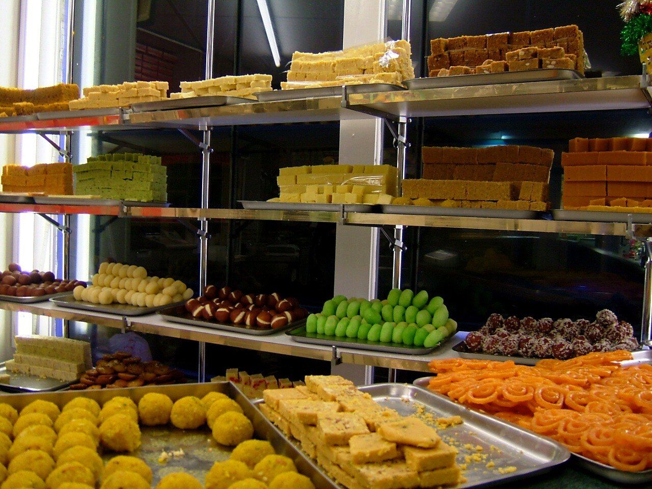 A sweet shop counter with steel trays of laddoos, barfi, jalebi, gulab jamun and other mithai on glass shelves