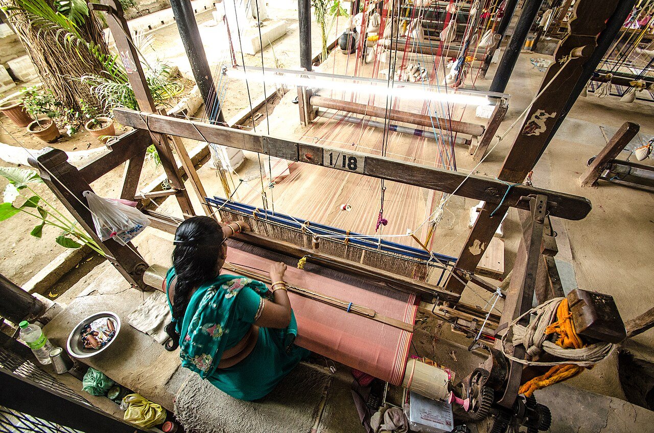 A woman in a green sari weaving at a wooden handloom in the weavers' workshop inside Maheshwar fort, seen from above and behind, with a length of pink cloth on the loom and more looms behind her