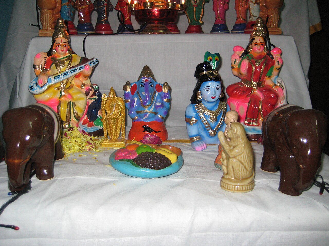 A Golu display on white-draped steps: painted clay dolls of Saraswati with a veena, Ganesha, Krishna and Lakshmi, a small gold Murugan, two glazed brown elephants and a plate of clay fruit, with a row of smaller dolls and a lamp on the step above