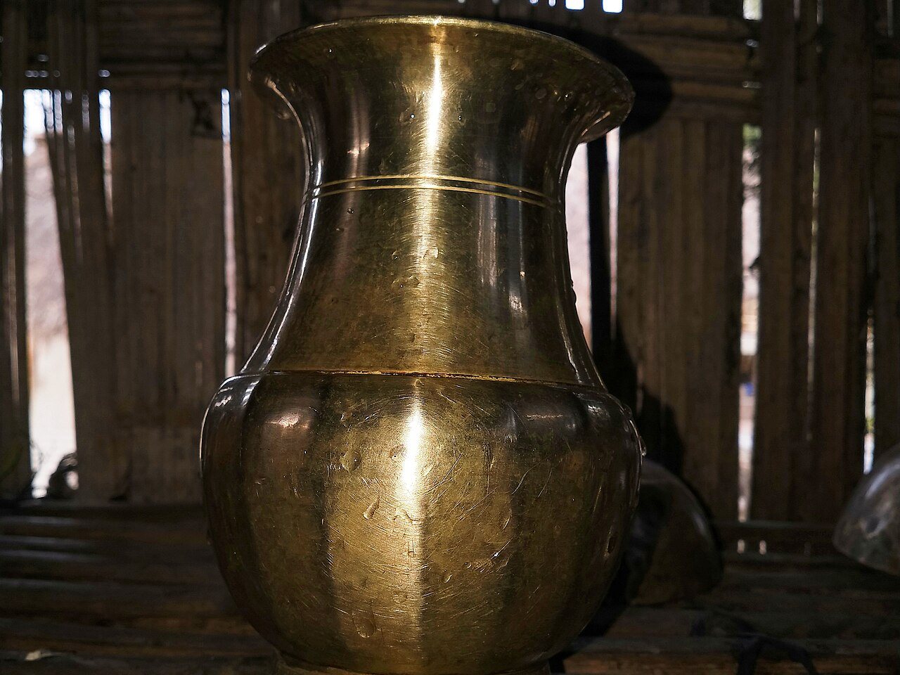 A traditional Assamese bell-metal ghoti, a water pot with a flared neck and a rounded, lobed body, standing on a bamboo slat shelf