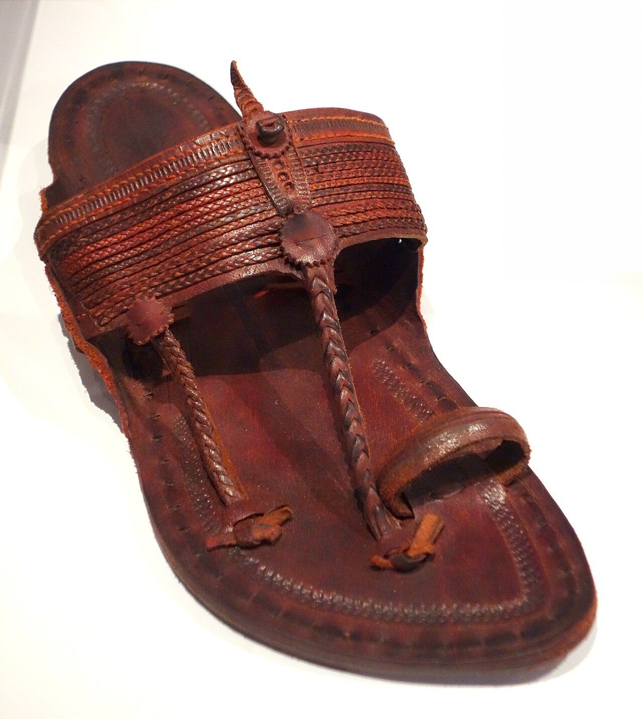 A single dark-brown Kolhapuri chappal with a braided toe strap, a broad braided band across the foot and a hand-stitched leather sole, on a white museum plinth
