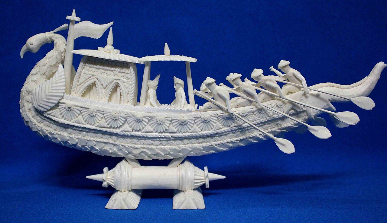 A carved white shola peacock boat on a blue cloth: a peacock-headed prow, a canopied cabin with two seated figures, a row of rowers with long oars and a tail curving up at the stern, set on a carved stand